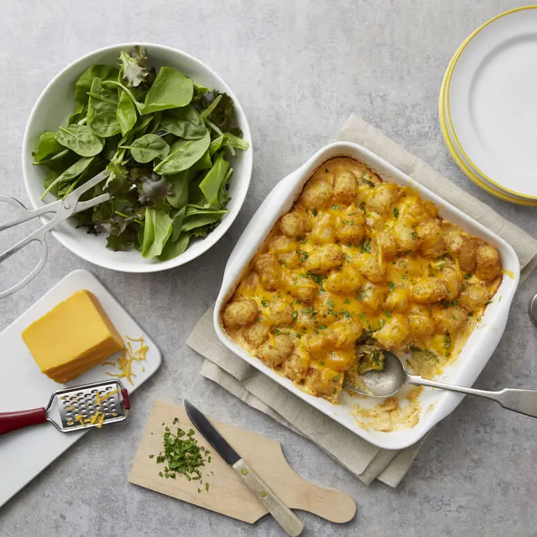 Recipe image of cheesy broccoli chicken potato casserole next to a bowl of salad, a block of cheese and a cutting board