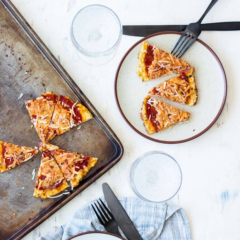 Sheet pan with five slices of mini cauliflower pizza next to a plate with three slices of pizza, for the recipe