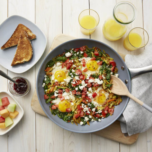 Recipe image of eggs florentine veggie hash skillet next to glasses of orange juice, slices of toast and a bowl of cut fruit