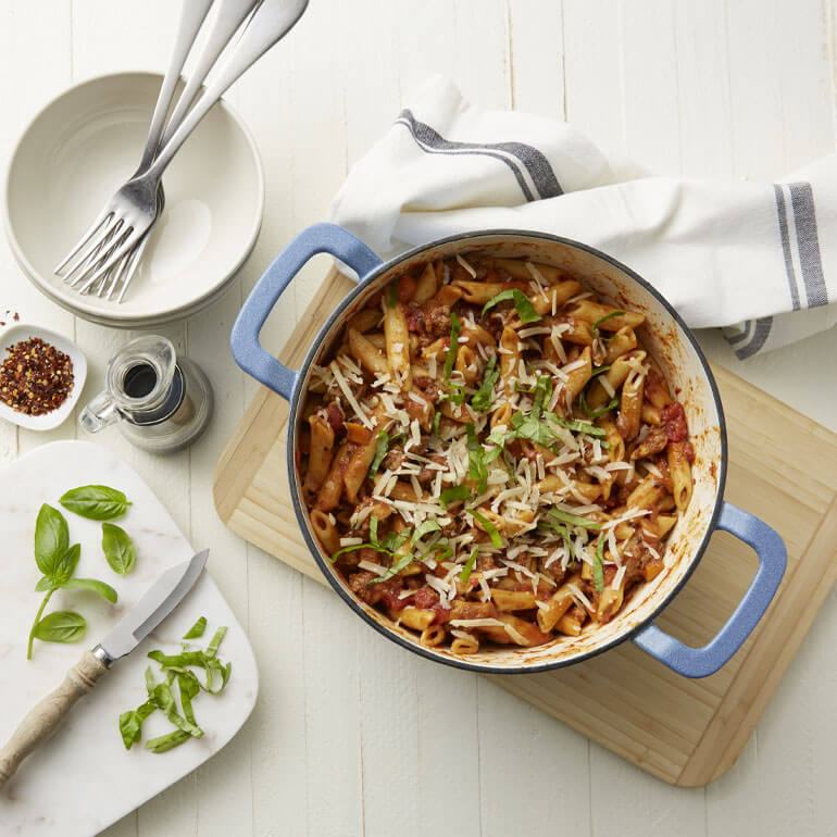 Recipe image of one-pot penne bolognese topped with basil next to empty bowls, a cutting board and crushed red pepper flakes